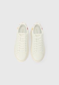 Pár bílých sneakers Guess s tkaničkami, logem na jazykách a jemnými barevnými akcenty v oblasti límců paty, zobrazené z vrchu.