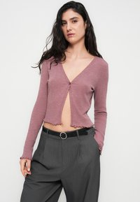 Πλεκτή, μωβ κοντή ζακέτα με ριμπ ύφανση, μακριά μανίκια, V-λαιμόκοψη και ακατέργαστο τελείωμα. Συνδυασμένη με γκρι ψηλόμεσα παντελόνια και μαύρη ζώνη με κυκλική αγκράφα.