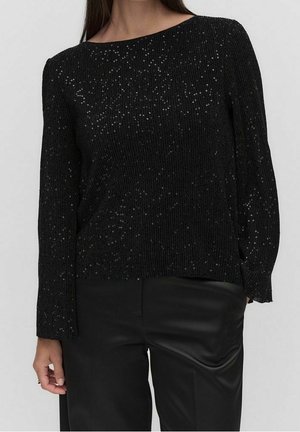 Woman wearing a black ribbed sweater with scattered sequins and black satin pants, standing against a plain light background.