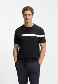 Schwarzes T-Shirt mit kurzen Ärmeln aus Baumwolle, mit einem horizontalen weißen Streifen und dem Schriftzug "BOSS" sowie einer glatten Textur. Kombiniert mit navyblauen Hosen.