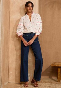 Blusa bianca con motivi di ciliegie rosse e maniche a volant, abbinata a jeans a vita alta blu scuro e sandali con tacco marroni, su uno sfondo neutro.