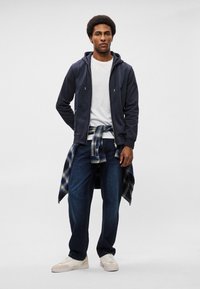 Mornarska zip-up dukserica preko bijele majice, uparena s tamnim trapercima. Plava i zelena karirana košulja zavezana u struku, bijele tenisice.