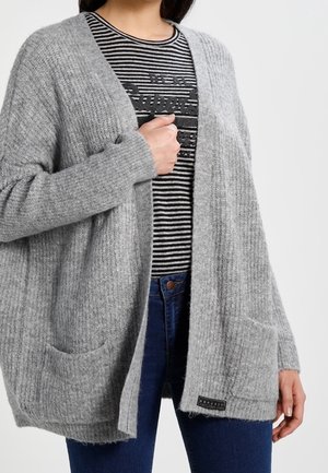 Femme portant un cardigan gris ouvert en maille par-dessus une chemise rayée noire et blanche et un jean bleu, tenant le cardigan d'une main.