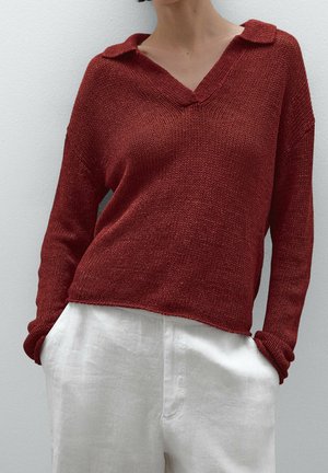 Person wearing a loose, long-sleeve rust-red knit sweater with a collar and white high-waisted linen pants, hands in pockets.