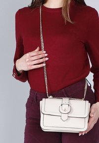 Donna con top e pantaloni bordeaux che tiene una borsa a tracolla in pelle avorio con dettagli in oro e fibbia.
