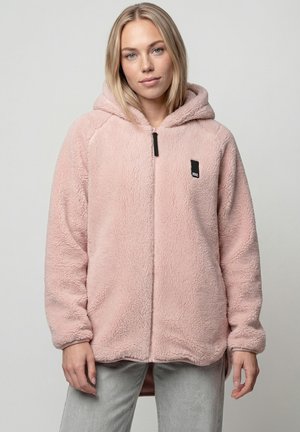 Junge Frau trägt eine hellrosa, flauschige Kapuzenjacke mit Reißverschluss und hellgraue Hose, steht vor einem schlichten hellen Hintergrund.