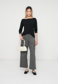 Jonge vrouw staat in een zwarte top en brede grijze broek, met een kleine witte handtas, en draagt zwarte flats voor een effen witte achtergrond.