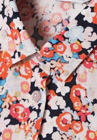 Blomsterknap-shirt med marineblå bund, flerfarvede blomster i rød, pink og blå, lavet af et glat stof og med pink knapper.