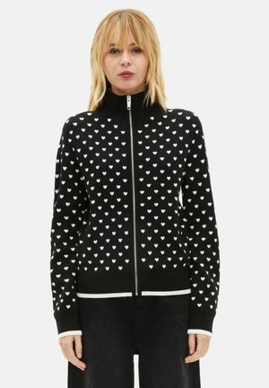 Zwarte vest met ritssluiting en wit hartjespatroon, hoge kraag, contrasterende manchetten en zoom, en een zachte stoftextuur.