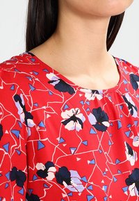 Blusa rossa con un motivo floreale nei toni del blu e bianco, scollatura rotonda e una texture di tessuto morbido.