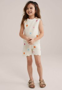 Completo a due pezzi lavorato a maglia in crema, composto da un top senza maniche e pantaloni corti a taglio scallop, decorato con colorati motivi floreali. Indossato con sandali marroni.