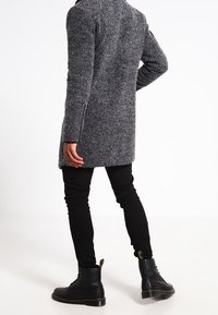 Abrigo de mezcla de lana gris con un ajuste entallado, mangas largas y superficie texturizada, combinado con jeans negros ajustados y botas negras de cordones.