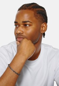 Bracciale in catena nera esposto su un polso marrone chiaro, indossato con una maglietta bianca semplice. La pelle ha una texture liscia.