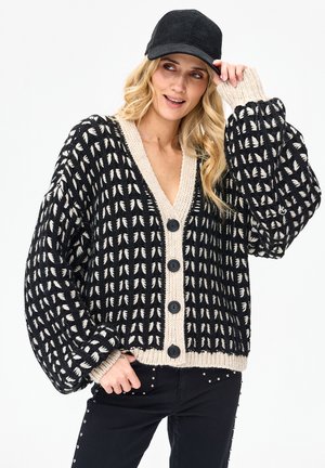 Kobieta w czarno-beżowym grubym swetrze z dużymi guzikami i czarnej czapce, uśmiechająca się i poprawiająca czapkę jedną ręką.