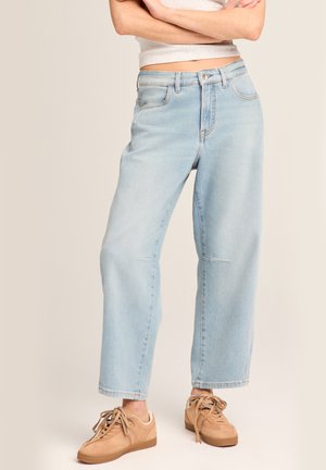 Person trägt hellblaue, hoch taillierte, weit geschnittene Jeans und beigen Sneaker, steht mit verschränkten Armen über dem Bauch vor beigem Hintergrund.