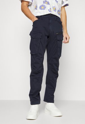Man draagt donkerblauwe cargobroek met meerdere zakken, gecombineerd met witte sneakers en een wit bloemenoverhemd met korte mouwen.