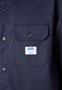 Mørkeblå skjorte med knapper, laget av teksturert stoff, med en brystlomme med en hvit etikett og svarte knapper.