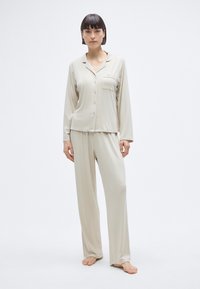 Σετ μπεζ πυζάμες από απαλό ύφασμα, που περιλαμβάνει μακρυμάνικο πουκάμισο με κουμπιά και τσέπη, καθώς και παντελόνι χαλαρής εφαρμογής που ταιριάζει.