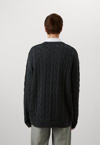 Dark gray cable-knit sweater with a ribbed crew neck and loose fit, featuring textured patterns and dropped shoulders, worn over light trousers.