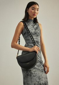 Frau in ärmellosem grauem Kleid hält eine schwarze, halbmondförmige Lederhandtasche mit breitem geflochtenem Schulterriemen vor einfachem Hintergrund.