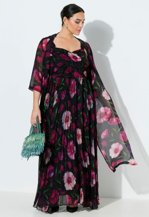 Kvinde iført en sort blomster-print plisseret maxi-kjole og matchende gennemsigtigt sjal, holdende en lille turkis frynset håndtaske, stående mod en hvid væg.