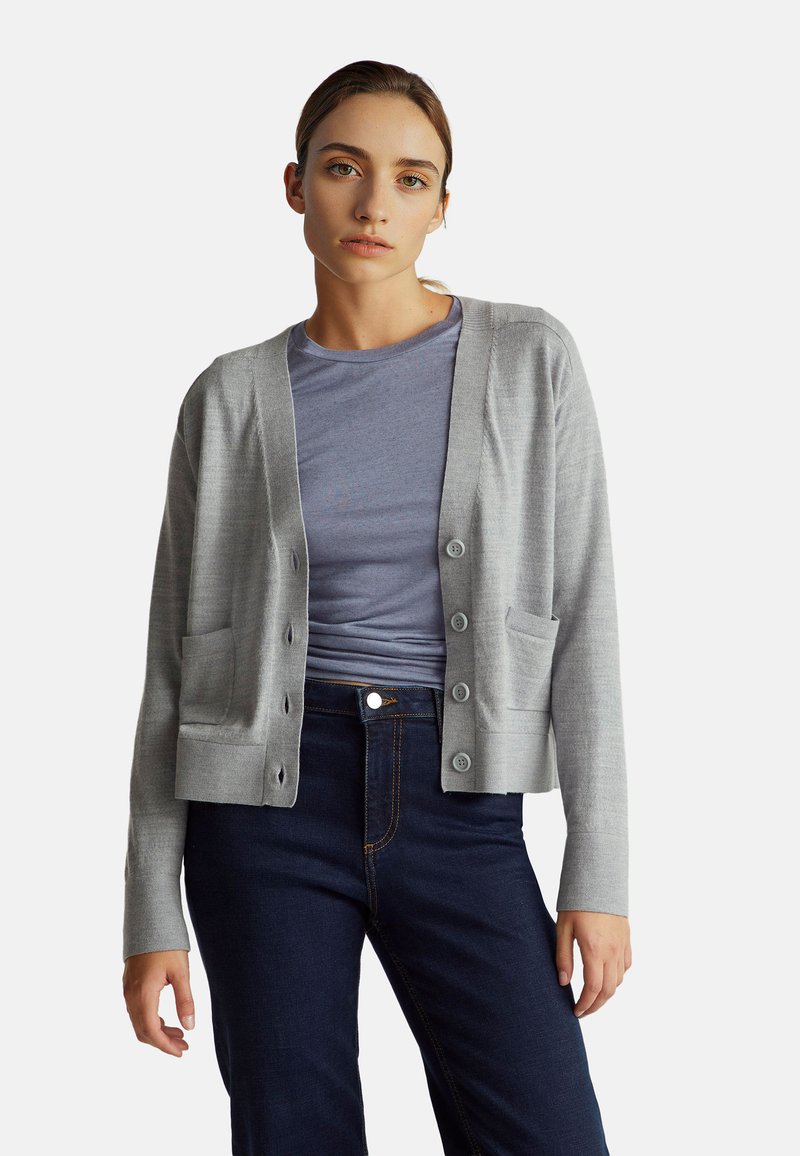 Cardigan gris avec des boutons sur le devant, décolleté en V et poches, associé à un t-shirt bleu, porté avec un jean sombre. Texture lisse, style ajusté.