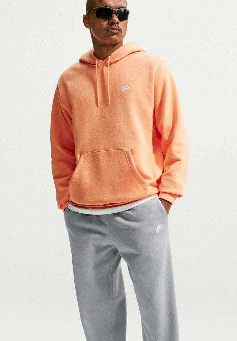 Mand iført sorte solbriller, en ferskenfarvet Nike-hættetrøje og grå Nike-sweatpants, stående med hænderne i lommerne.