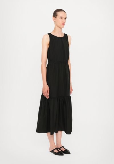 Black sleeveless dress with a gathered neckline, waistline seam, and tiered skirt. Smooth texture and knee-length design. Accented with flat black shoes.