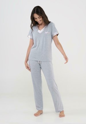 Joven mujer descalza con un conjunto de pijama gris claro con ribete de satén, de pie mirando hacia abajo con una ligera sonrisa sobre un fondo sencillo.