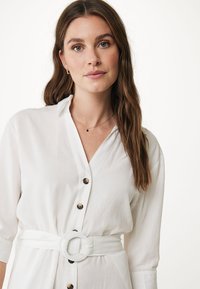 Camicia bianca con bottoni, maniche lunghe, dotata di una cintura in vita con una fibbia circolare e dettagli in bottone marrone. Tessuto dalla consistenza liscia.