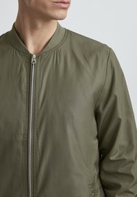 Olivengrüne Bomberjacke aus glattem Nylon, mit Reißverschluss vorne, geripptem Kragen und Seiten Taschen im minimalistischen Design.