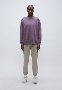 Μωβ φούτερ με ριμπ μανσέτες και τελείωμα, με μικρό λογότυπο. Συνδυασμένο με ανοιχτό μπεζ φόρμες και λευκά αθλητικά παπούτσια.