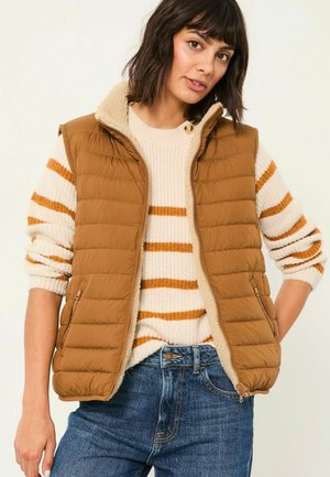 Braune Steppweste mit gestepptem Design und Sherpa-Kragen, getragen über einem cremfarbenen Pullover mit orangefarbenen Streifen. Kombiniert mit blauen Denim-Jeans.