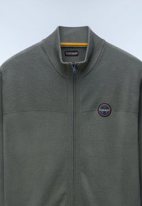 Olivgrön zip-up tröja med ett texturerat övre panel, ribbat krage och en rund logobadge på bröstet. Slät stickad tyg överlag.