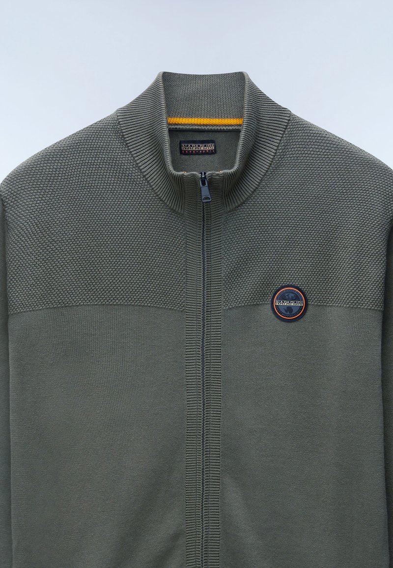 Sweat à fermeture éclair vert olive avec un panneau supérieur texturé, un col en côtes et un badge logo rond sur la poitrine. Tissu tricoté lisse dans l'ensemble.