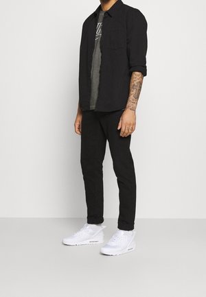 Chemise noire à boutons sur un t-shirt gris, assortie à un pantalon noir et des baskets blanches. Les caractéristiques incluent des manches retroussées et des tatouages visibles.