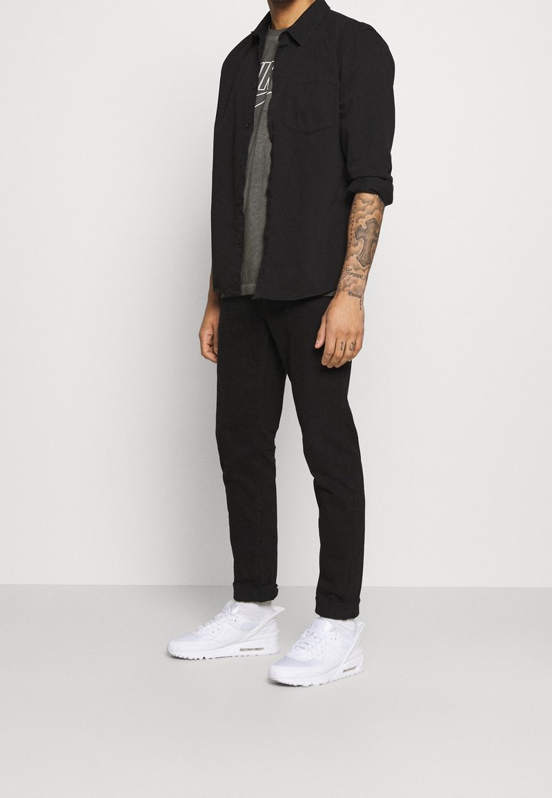 Svart skjorta med knappar över en grå t-shirt, i kombination med svarta byxor och vita sneakers. Funktioner inkluderar upprullade ärmar och synliga tatueringar.