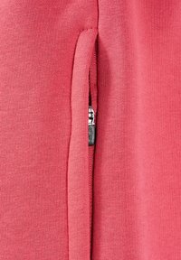 Gros plan d'une poche verticale partiellement fermée par une fermeture éclair argentée visible sur un tissu en maille rose.