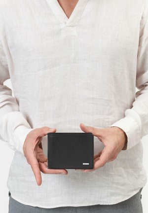 Billetera de cuero negro con superficie texturizada, forma rectangular y un acento de logo plateado, sostenida en manos contra un fondo claro.