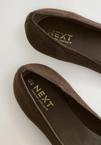 Pruunist nahast balleti kingad ümarate varvastega, elastse äärisega ja kullatud "NEXT" brändinguga sees. Pehme tekstuur ja mugav klapp.