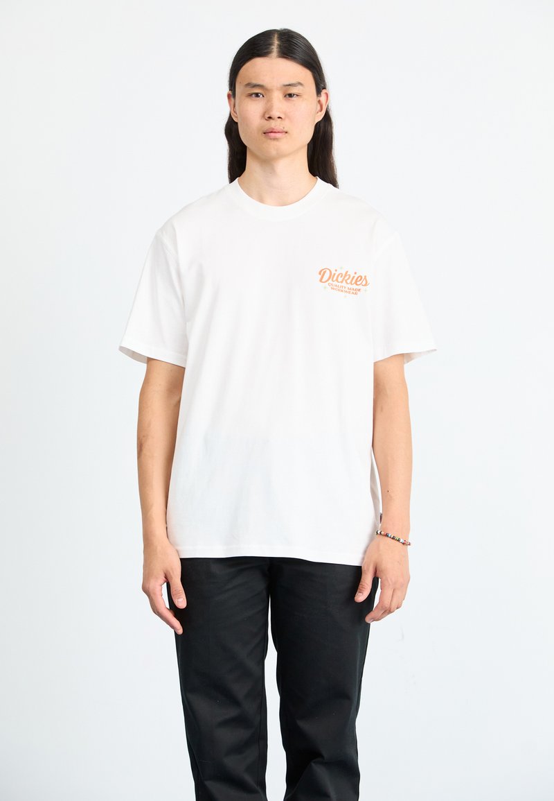 Witte katoenen t-shirt met korte mouwen, met daarop oranje bedrukt tekst "Dickies" op de linker borst. Gedorven met zwarte broek.