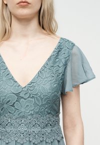 Prim-plan cu o femeie care poartă o rochie albastră din dantelă cu modele florale și mâneci transparente evazate, evidențiind decolteul și partea superioară a trunchiului.