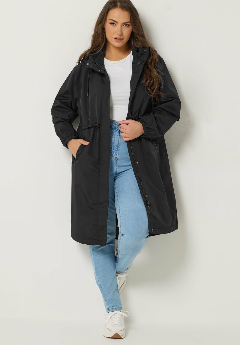 Kvinde iført sort lang hættejakke, hvid skjorte, lyseblå jeans og hvide sneakers, stående mod en ensfarvet lys væg.