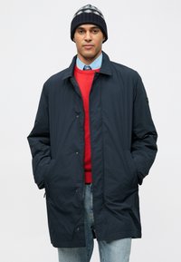 Muž v námorníckom modrom kabáte, červenom svetri, svetlomodrej košeli s gombíkmi, pletenej čiapke a džínoch, s rukami v vreckách kabátu, na jednoduchom pozadí.