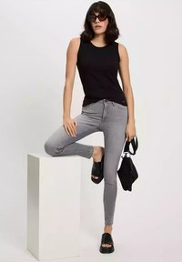 Czarna bezrękawnik, szare dopasowane dżinsy, czarne platformy na sandałach i mała czarna torba. Modelka pozując na białym cokole na neutralnym tle.