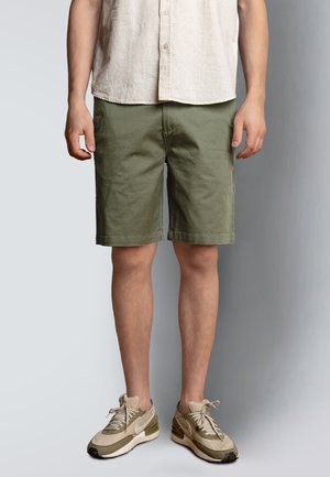 Hombre con camisa beige de botones, pantalones cortos verde oliva y zapatillas beige, posando contra un fondo gris liso.