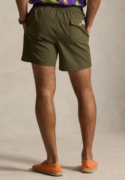 Άνδρας που φοράει χακί σορτς με πίσω τσέπη και πορτοκαλί παντόφλες, στέκεται σε ουδέτερο φόντο.