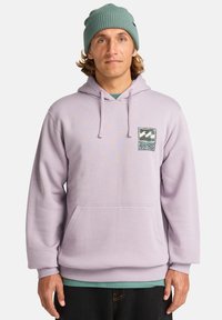 Felpa viola chiaro con tasca frontale, cappuccio con coulisse e patch con logo. Abbinata a una maglietta verde sotto e pantaloni neri.