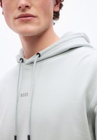 Κοντινό πλάνο ενός ατόμου που φοράει ανοιχτό γκρι φούτερ με κορδόνια και μικρό λογότυπο "BOSS" στο στήθος.