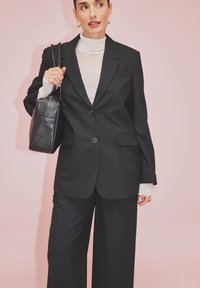 Must värvitud bleiser ühe nupuga, koos laia jalgadega mustade pükstega. Kerge turtleneck ja must käekott lõpetavad välimuse.